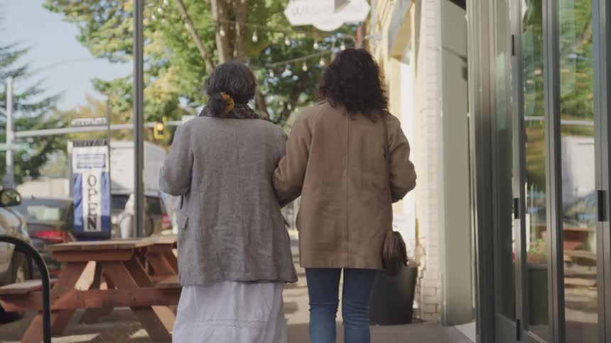 Two women walk arm in arm down a sidewalk in a small town on a sunny day