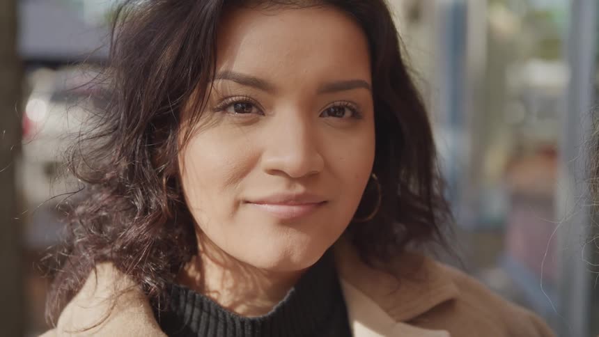 A young woman looks calmly into the camera while standing outside in natural light