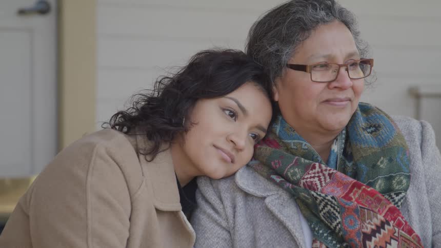 Two women sit closely together one resting their head on the others shoulder both wearing coats with one in a colorful patterned scarf