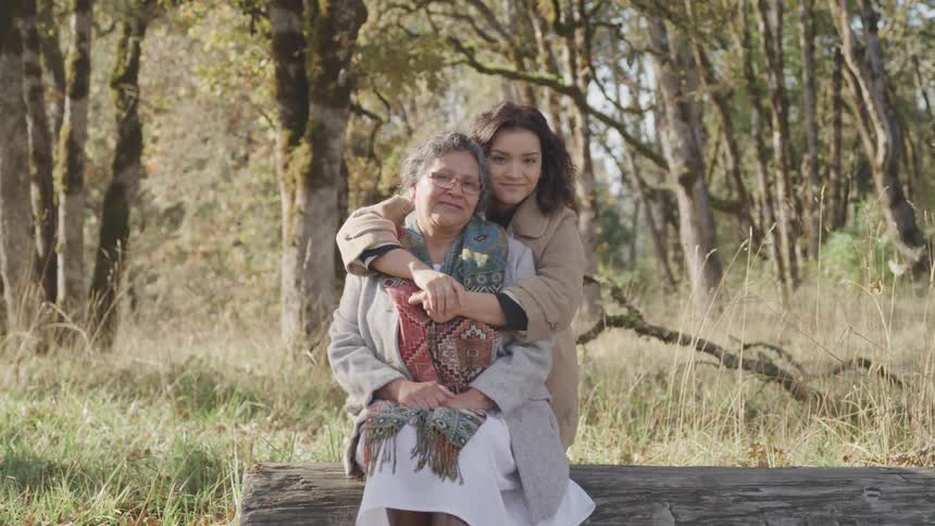 A daughter lovingly hugs mother from behind as they sit on a bench in a sunlit forest