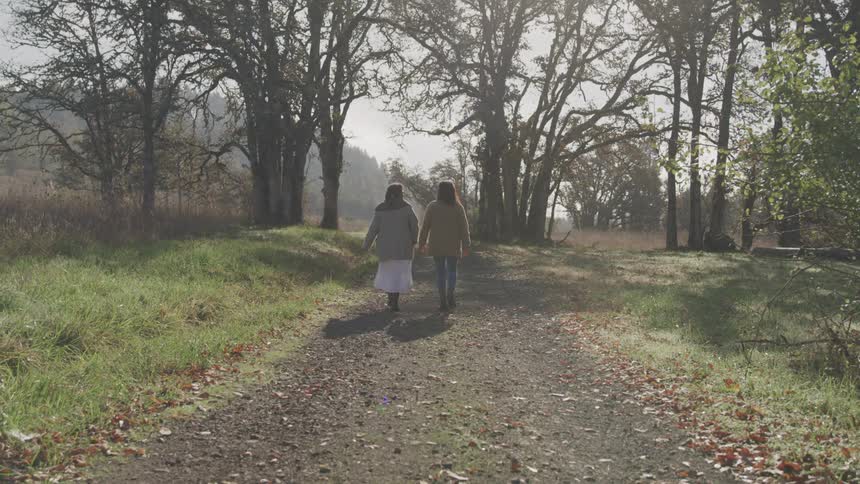 Two girls walk hand in hand along a dirt path flanked by tall grass and trees under bright sunlight