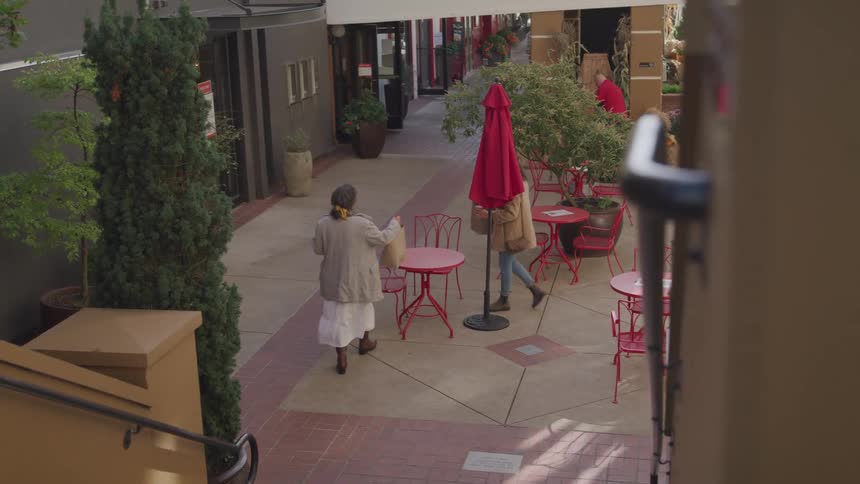 A young woman and an elderly woman walk up to a coffee table on the sidewalk and set up their items on top of it around midday