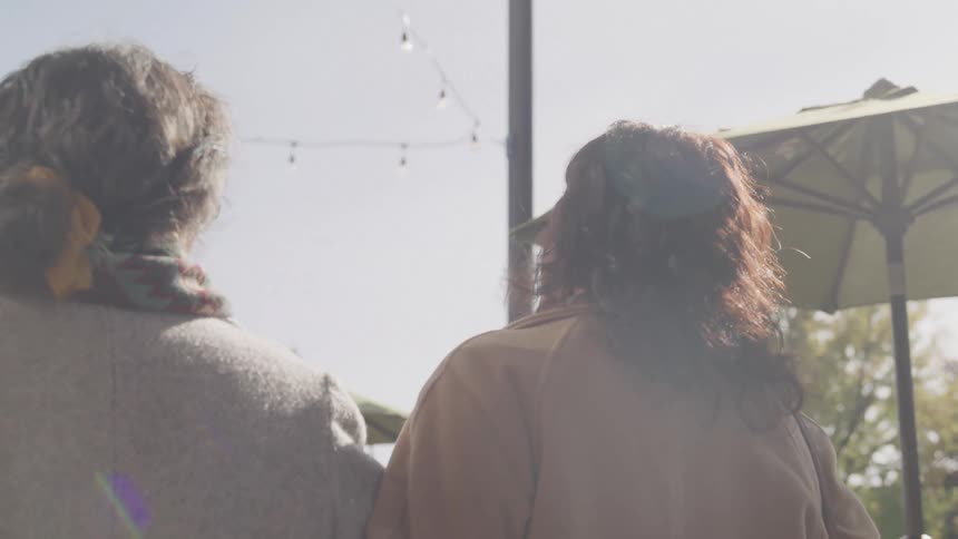Two women with curly hair walk down a street under string lights near large umbrellas seen from behind