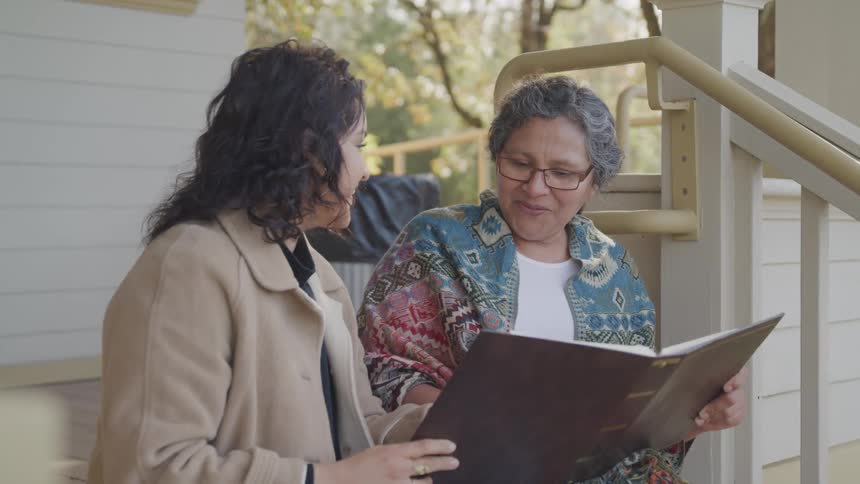 Two women one older than the other sitting on the stairs on the left side of the house and looking at an album