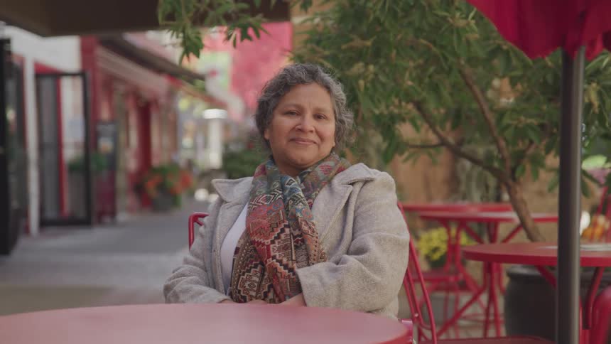 A woman sits alone at a red table in an outdoor caf with matching chairs surrounded by trees and colorful buildings