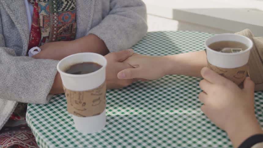 Two people sit across a table holding hands in a warm comforting moment over coffee