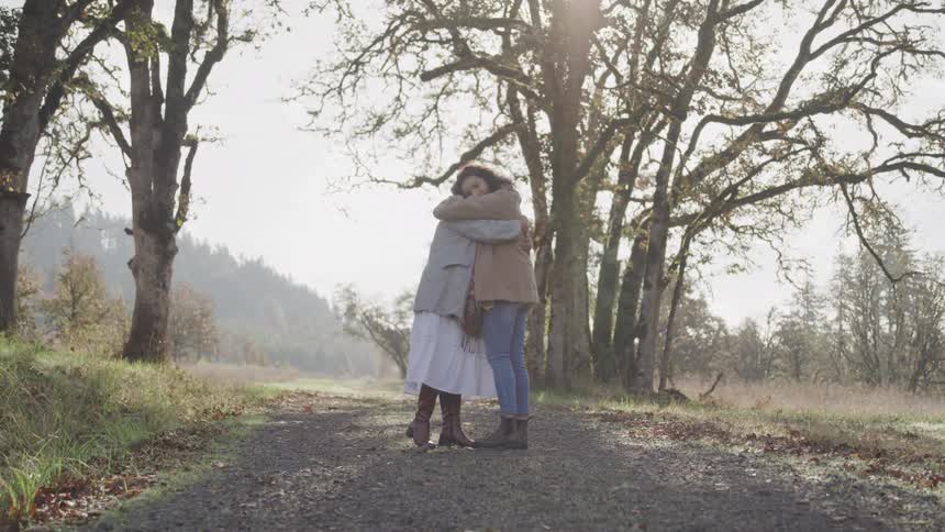 Two women embrace on a sunlit gravel path surrounded by tall trees with rays of light filtering through the canopy