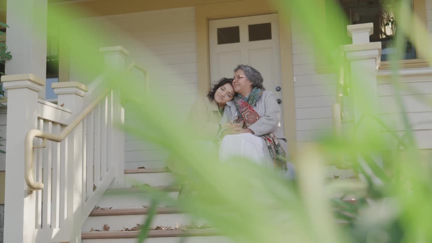 An elderly mother and her daughter sit side by side on the front porch of a suburban home as the daughter hugs her mother with smiles on their faces