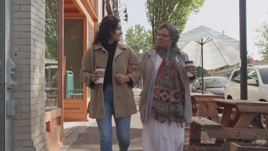 Two women walk together on a city sidewalk holding coffee cups and smiling