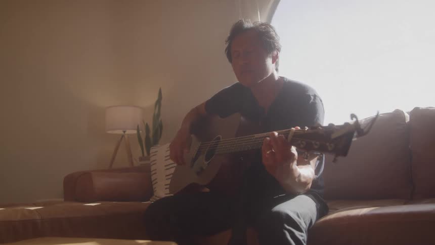 Older musician plays a guitar while sitting on a couch near a window
