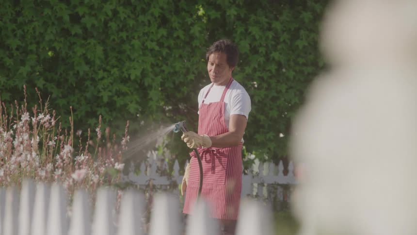 A gardener in a red apron waters flowers in a sunny garden by a white picket fence