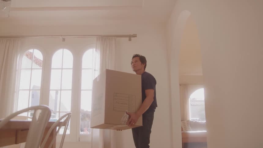 Man carrying a large cardboard box inside a sunlit room