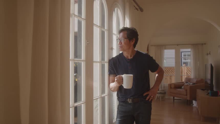 A man looking out the window while holding a coffee mug in the morning