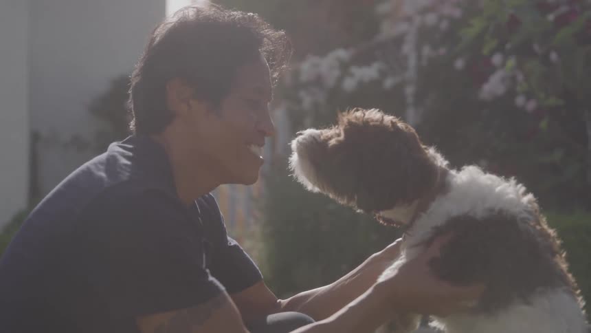 A person holds a dog close as it licks his face in a garden filled with blooming flowers and dense foliage