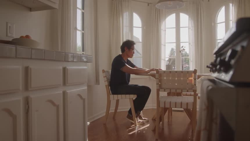 A man sits alone at a dining table in a sunlit kitchen working on a laptop