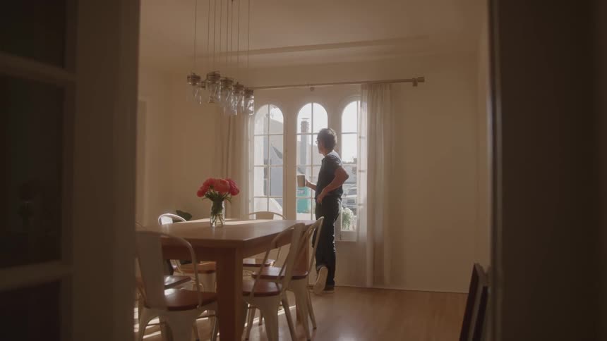 A man stands by a large arched window in a sunlit room holding a mug with a dining table and flowers in the foreground