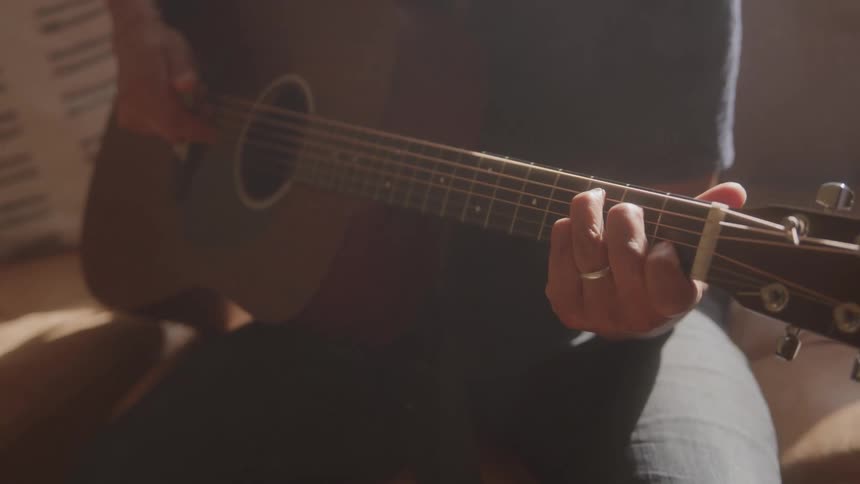 A person sits on a couch playing an acoustic guitar in a room bathed in warm sunlight