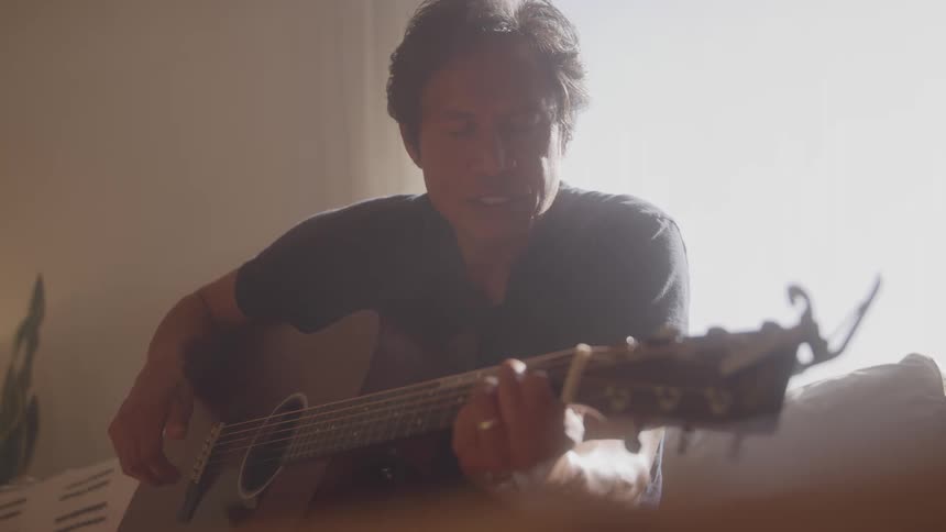 A man sits indoors playing an acoustic guitar with focused expression