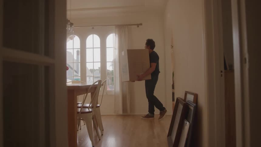 A man carries a large cardboard box through a sunlit room with wooden floors and tall arched windows