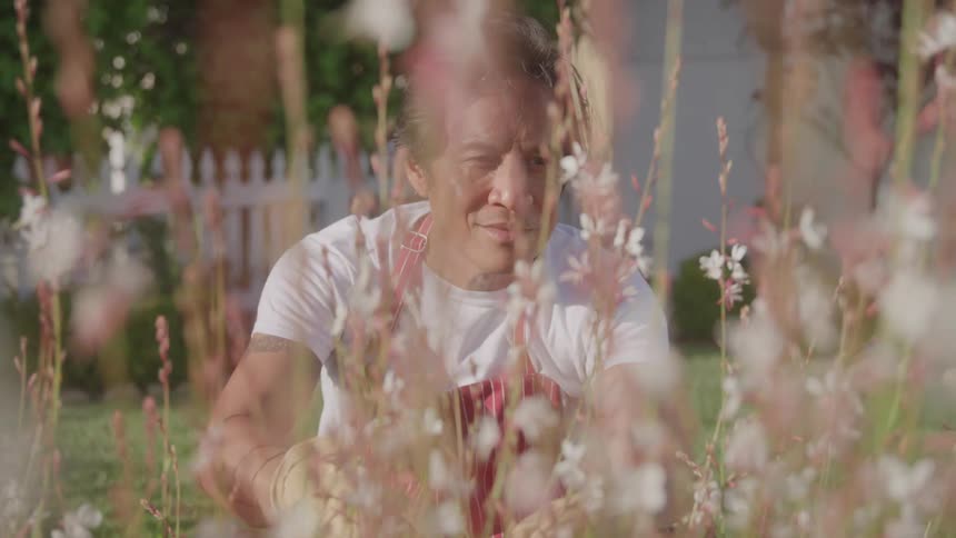 A man in a white tshirt and red apron tending to his garden in the backyard