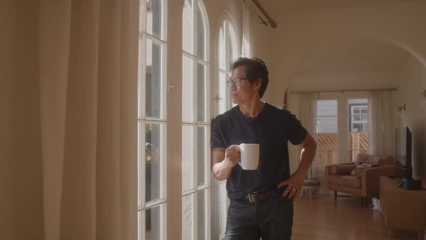 A man holding a mug looks out a tall window lost in thought inside a sunlit home