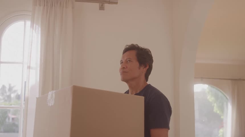 A man carrying a box stands beside a window inside a house looking left and right with a smile on his face around midday