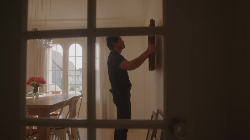 A man picks up a frame from the floor of his dining room and hangs it on the wall around midday