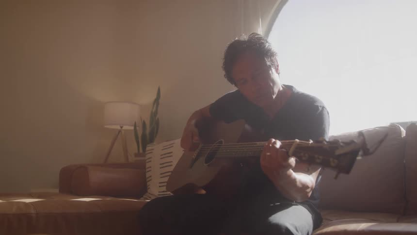 Man sitting on a couch playing acoustic guitar in a warmly lit living room