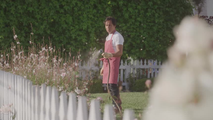 A person in a red apron waters plants beside a white picket fence in a garden with dense foliage