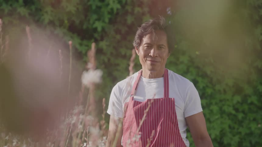 A gardener wearing an apron stands in the garden around midday with a smile