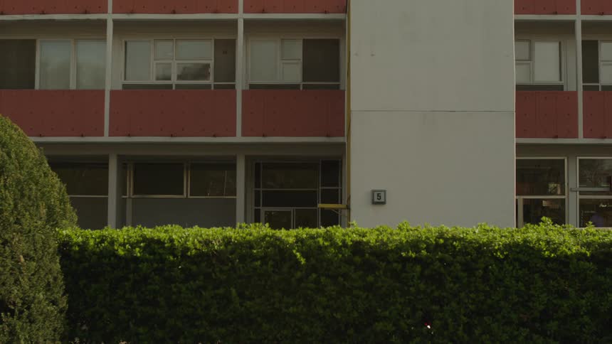 A midcentury apartment building is partially obscured by a neatly trimmed hedge in front