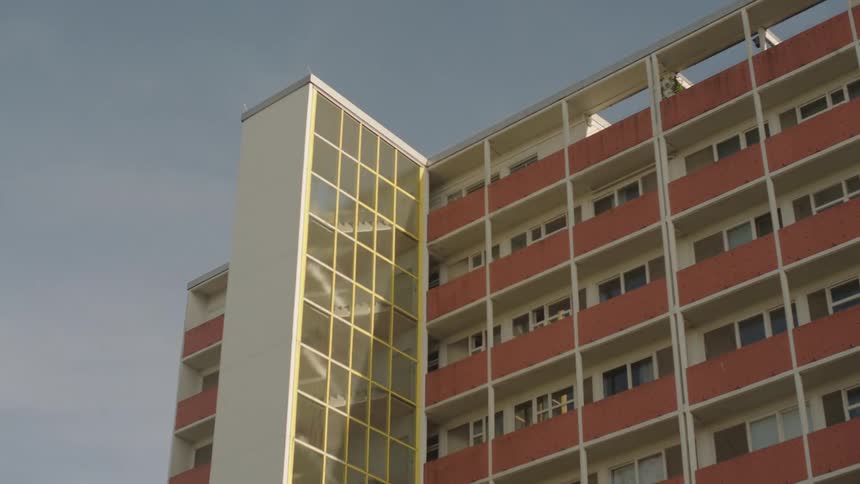 A multistory apartment building with red and white balconies is captured from below against a clear sky