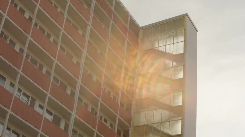 Sunlight flares off the glass stairwell of a large apartment building highlighting its repeating facade of brick and balconies