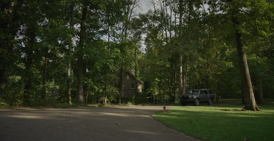 A gray suv drives on a paved driveway surrounded by tall green trees and a grassy lawn with a small house in the background