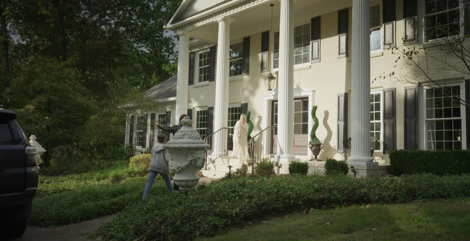A family of three approach the front entrance of a large white house with columns surrounded by manicured landscaping and trees