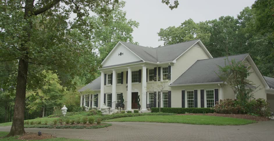 A large elegant suburban house is surrounded by lush greenery on a quiet day