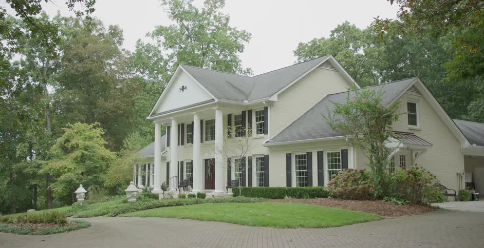 A large elegant house with white pillars and black shutters sits surrounded by trees and landscaping