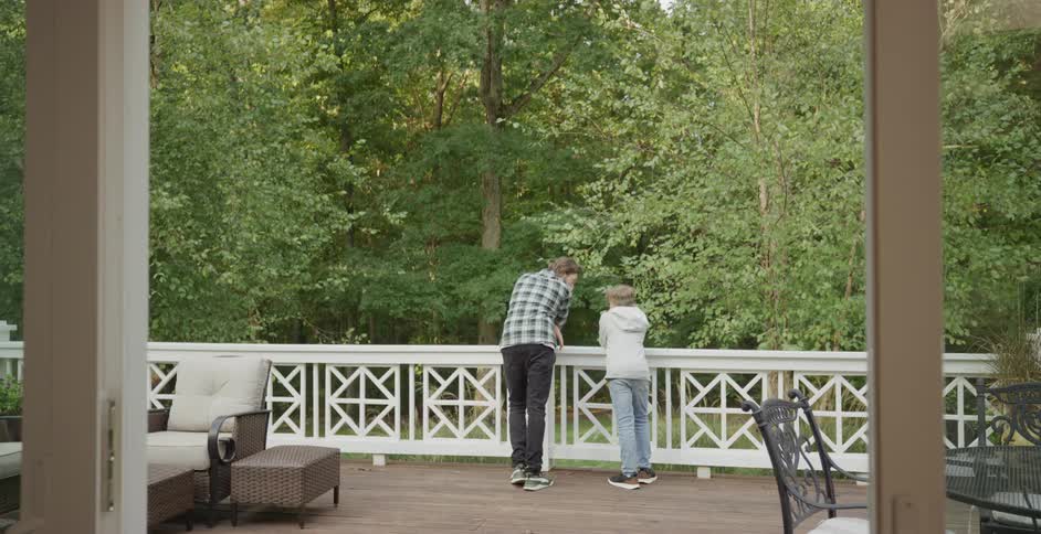 Father and son lean against a white railing on a wooden deck looking out toward a dense green forest