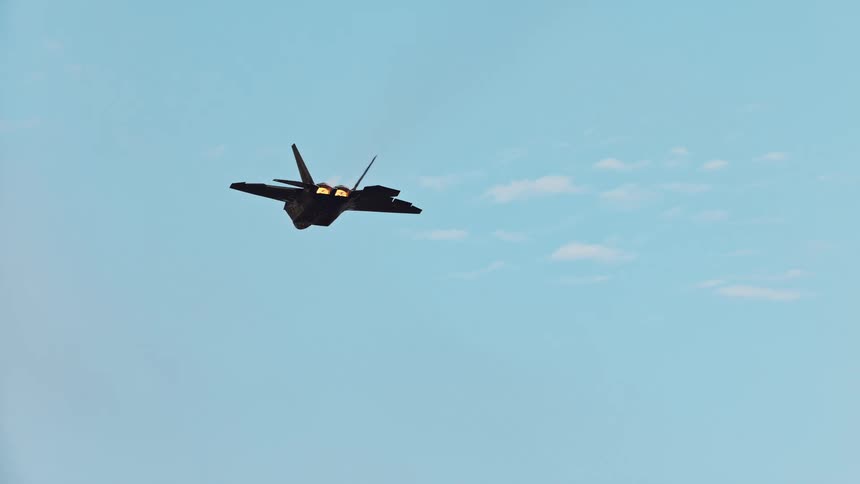 A dark fighter jet with afterburners glowing and contrails streaming from its wings flies across a bright blue sky
