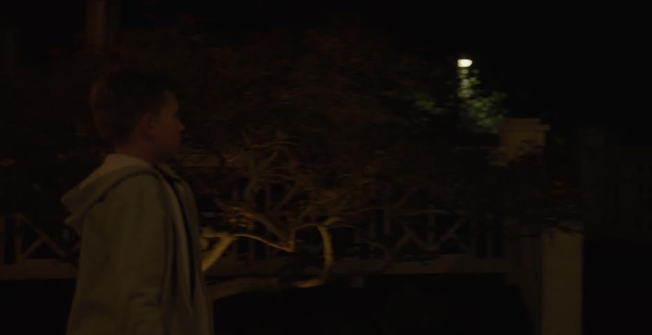 A boy places a large garbage bag into a trash bin beside a white wall outside a house at night
