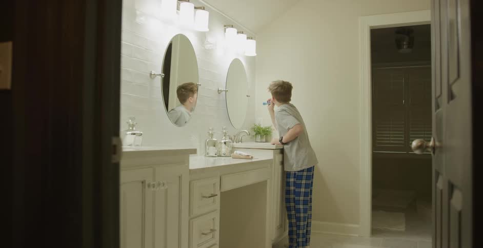 A young boy is brushing his teeth while standing in front of a bathroom mirror
