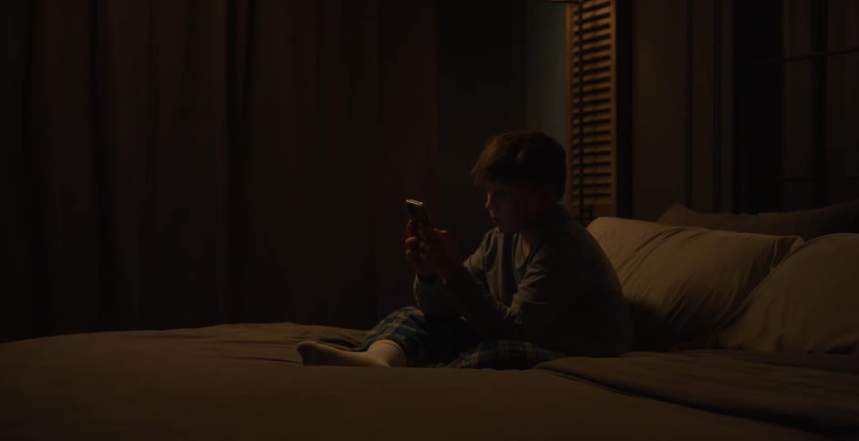 A child sits alone on a bed at night looking at a smartphone