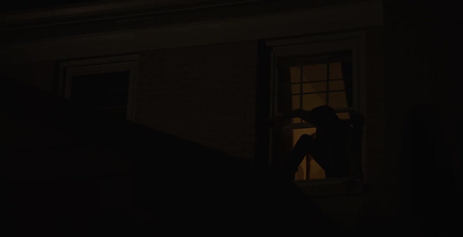 A silhouetted teenager steps out through a lit window onto the roof of a house at night