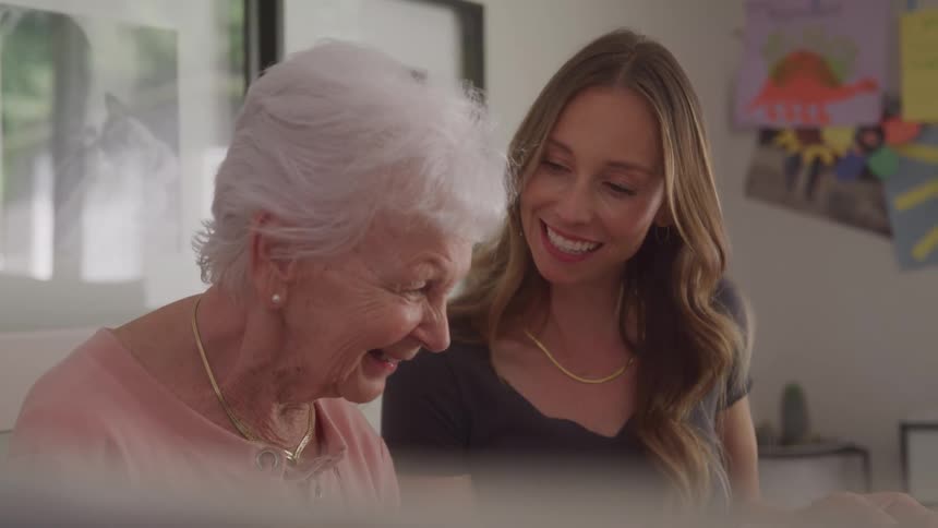 A young woman and an elderly woman sit on the couch around midday going through a magazine with smiles on their faces