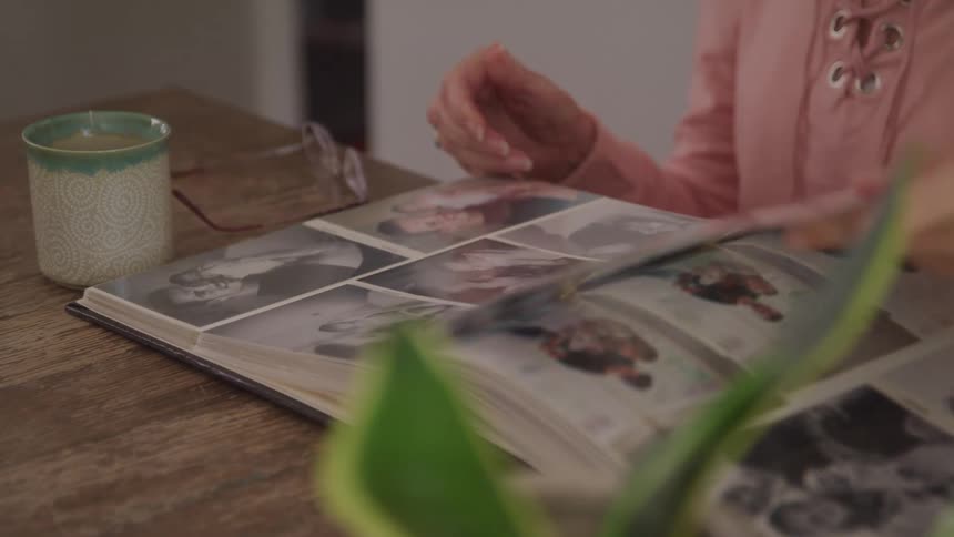 An elderly woman flips through a photo album at a wooden table reflecting on memories