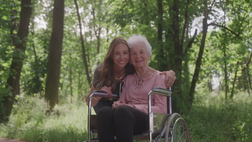 Young woman poses with an elderly woman in a wheelchair outdoors in a woodland area during daytime