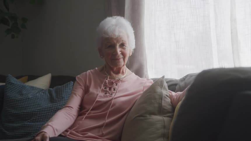 An elderly woman with short white hair wearing a pink top and necklace sits on a couch with pillows and looks directly at the viewer