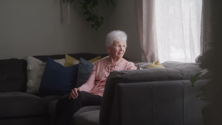 An elderly woman with white hair sits thoughtfully on a sofa looking out a window