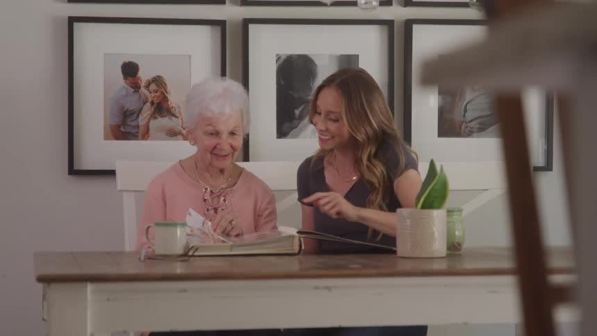 An elderly woman and a younger woman sit at a table reading a book together