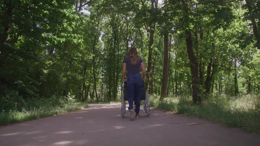 A woman walks down a forest path while pushing someone in a wheelchair from behind